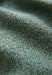 Strukturierter Stoff in sanftem Grün mit einem subtile Wellenmuster. Das Material wirkt weich und leicht, mit einer gewellten Oberflächendetail.