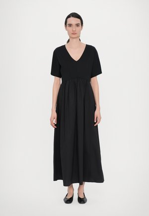 Robe maxi en coton noir avec un décolleté en V, des manches courtes et une taille froncée, présentant une texture lisse et une coupe décontractée, associée à des ballerines noires.