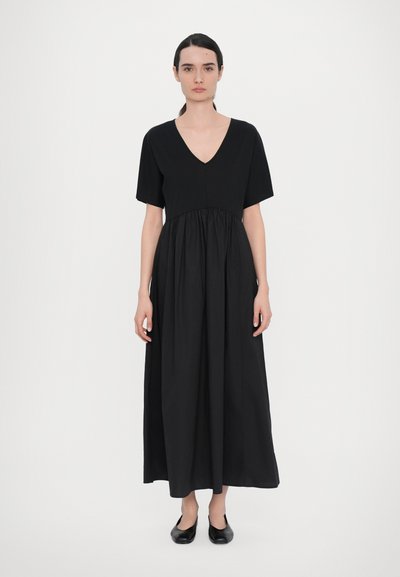 Abito maxi in cotone nero con scollatura a V, maniche corte e vita arricciata, caratterizzato da una texture liscia e una vestibilità rilassata, abbinato a ballerine nere.