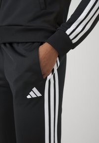Osoba nosząca czarne spodnie dresowe Adidas z białymi paskami i logo, ręka spoczywa w kieszeni.