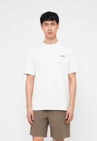 Νέος άνδρας με κοντά μαύρα μαλλιά, ντυμένος με λευκό T-shirt με μικρό κείμενο και καφέ cargo σορτς, στέκεται μπροστά σε απλό λευκό φόντο.