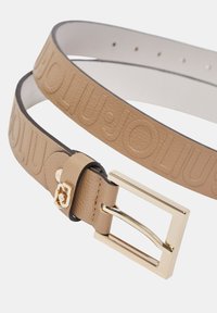 Ceinture en cuir beige avec nom de marque embossé, boucle dorée et doublure intérieure lisse. Réglable grâce à plusieurs trous pour un ajustement optimal.