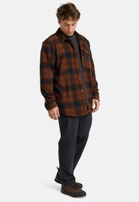 Karierter Flanellhemd in Braun und Schwarz mit Knopfleiste, einer einzelnen Brusttasche und einem lockeren Schnitt, kombiniert mit schwarzen Hosen und braunen Stiefeln.