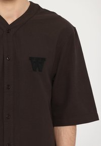 Dark brown short-sleeve shirt with a textured fabric, featuring a button-up front and an embroidered black "W" logo on the left chest.
