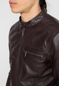 Hnědá kožená bunda s vysokým límcem, zipem vpředu a dekorativní kapsou se zipem. Vyznačuje se hladkou texturou a jemnými švy.