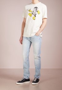 Ljust grå t-shirt med en karikatyr av en apa i svart och gult, kombinerad med ljusblå denimjeans och svarta sneakers.