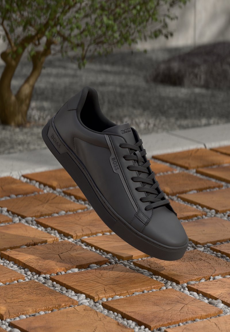 Baskets BOSS en cuir noir avec lacets, flottant au-dessus d’un bloc de bois et d’une surface extérieure en galets, avec un arbre flou et un fond de gravier.