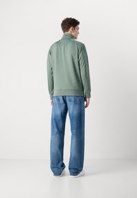 Grønn zip-up hettejakke med teksturert stoff, høy krage og ribbestrikkede mansjetter, kombinert med løse blå jeans og hvite sneakers.