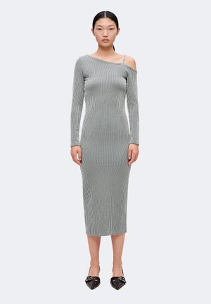 Femme portant une robe midi grise côtelée, une épaule nue, manches longues et chaussures noires à bouts pointus avec boucles, debout sur un fond uni.
