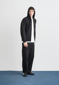 Felpa nera con zip e cappuccio, t-shirt bianca e pantaloni neri a gamba larga. La modella indossa scarpe nere con suola testurizzata su un tappeto blu.