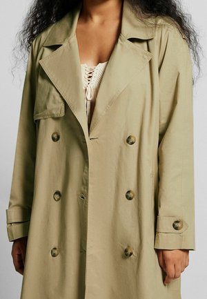 Kvinde iført lang beige trenchcoat med dobbeltradede knapper over en cremefarvet snørelukningstop, håret løst og krøllet, stående foran en ensfarvet baggrund.