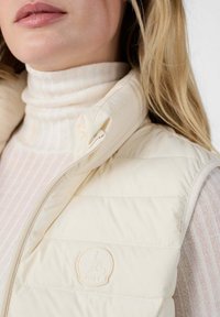 Gilet puffo di colore crema con colletto alto, zip sul lato e un patch con logo ricamato. Sotto, una maglia a collo alto a righe chiare.