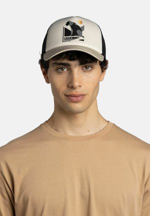 Joven con cabello rizado oscuro que lleva una gorra beige y negra con un gráfico de surfero y una camiseta beige de cuello redondo, fondo neutro.