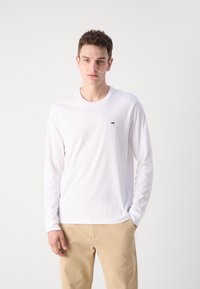 Camisa blanca de manga larga con cuello redondo y un pequeño logo. Combinada con unos pantalones beige claros, que tienen una textura de tela suave.