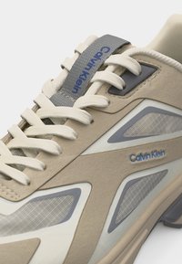Μπεζ και γκρι αθλητικό παπούτσι Calvin Klein με υφασμάτινα πάνελ, λευκά κορδόνια και μπλε λογότυπο στο πλάι και στη γλώσσα, σε κοντινή λήψη.