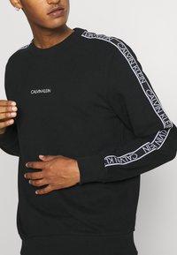 Felpa nera con collo a giro, caratterizzata dal logo bianco "CALVIN KLEIN" sul petto e lungo ciascun maniche. Polsini e orlo a coste. Texture di tessuto morbida.