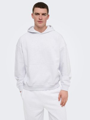 Jonge man die een effen lichtgrijze hooded sweatshirt en bijpassende lichtgrijze joggingbroek draagt, staand tegen een effen lichte achtergrond.
