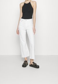 Schwarzes Neckholder-Top, hochgeschnittene weiße Jeans mit cropped, ausgestellten Beinen, schwarze Sandalen mit goldenen Hardware-Akzenten, glattes Material, minimalistisches Design.
