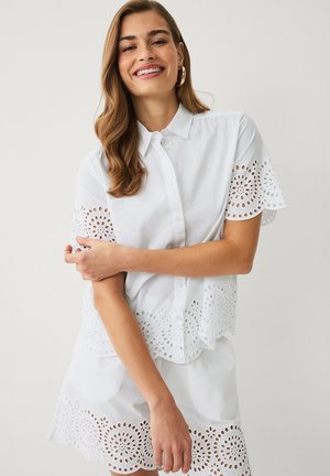 Smilende kvinde med langt hår iført hvid kortærmet skjorte med knapper og matchende nederdel med cirkulære udskårne blomster mønstre.