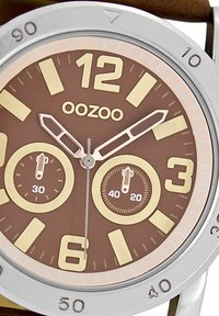 Cadran de montre marron avec des index horaires en or et une lunette en argent. Présente deux sous-cadrans et un bracelet en cuir marron. Logo OOZOO affiché.