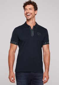 Granatowa koszulka polo z zamkiem przy kołnierzyku, krótki rękaw z szarymi akcentami oraz tekstem markowym na rękawach. Teksturowana tkanina dodaje szczegółów.