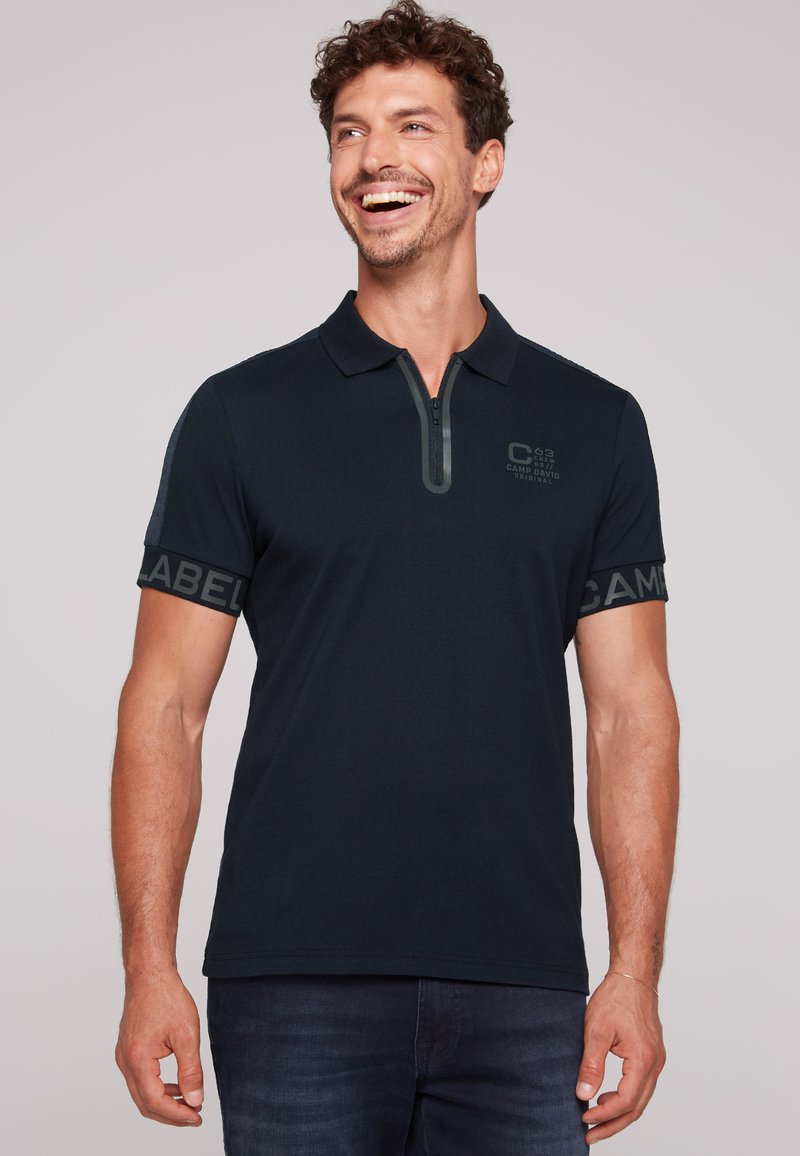 Granatowa koszulka polo z zamkiem przy kołnierzyku, krótki rękaw z szarymi akcentami oraz tekstem markowym na rękawach. Teksturowana tkanina dodaje szczegółów.