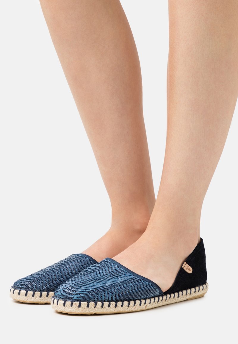 Pieds portant des chaussures bleu tissées à enfiler avec des semelles beige cousues sur un fond clair uni.