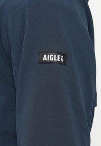 Σκούρο μπλε ύφασμα με ένα ραμμένο ορθογώνιο μαύρο ετικέτα που δείχνει λευκό κείμενο "AIGLE 1853" σε μανίκι.