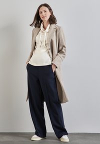Beige wool coat, cream cowl-neck hoodie, navy wide-leg trousers, and white sneakers with red accent. Smooth textures and tailored design.