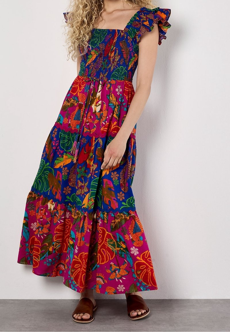 Femme portant une robe maxi colorée, à motifs, à volants avec des manches froncées et des sandales marron à bout ouvert, debout devant un mur blanc uni.