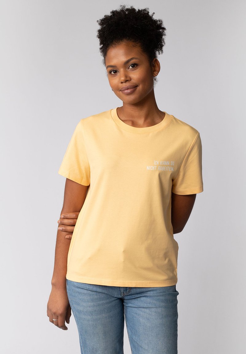 Geel katoenen T-shirt met korte mouwen, ronde hals en witte tekst "ICH KANN SO NICHT ARBEITEN" op de voorkant, gecombineerd met een blauwe spijkerbroek.