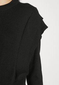 Pull en maille noire avec un col rond haut et des détails volantés le long de la couture de l'épaule sur une personne.