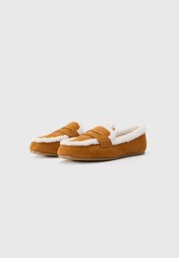 Tan suede slippers with white fleece lining, rounded toe, and a decorative strap across the upper. Flat sole for indoor use.