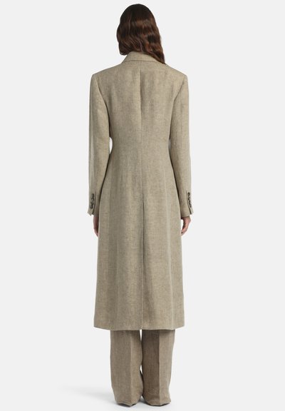 Femme aux cheveux longs et ondulés, portant un long manteau beige et un pantalon à jambes larges assorti, debout de dos sur un fond uni.