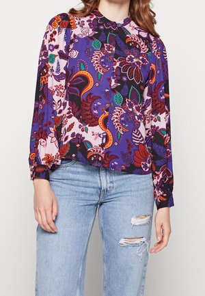 Femme portant un chemisier violet à motif floral coloré à manches longues, associé à un jean bleu clair déchiré.