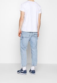 Man med vit t-shirt, ljusblå jeans, vita strumpor och marinblå Reebok-sneakers som står vänd bort från kameran mot en enfärgad bakgrund.