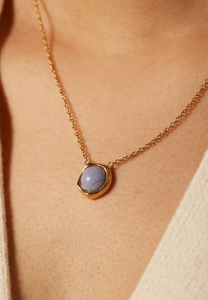 Gouden ketting met een ronde, lichtblauwe steen in een gestructureerde, ovale zetting. De hanger rust op een lichte huidskleur.