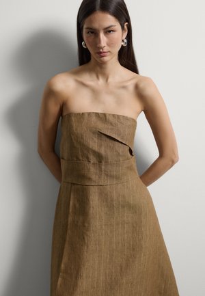 Vrouw met lang donker haar draagt een strapless bruine linnen jurk en zilveren oorringen, staand tegen een eenvoudige lichte muur.