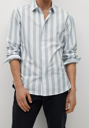 Homme portant une chemise à manches longues à rayures verticales bleu clair et blanches, avec les manches retroussées, assortie à un pantalon foncé.