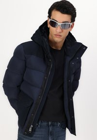 Mornarska puffasta jakna s kapuco, z zadrgo spredaj, s snap gumbi in zunanjimi žepi. Nošena preko črne majice in denim kavbojk.
