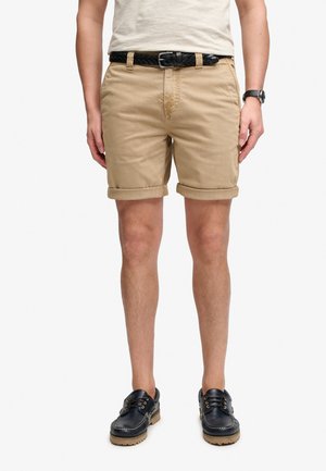 Mężczyzna ubrany w beżowe szorty podwinięte, czarny pleciony pasek, białą koszulkę, czarny zegarek na rękę i czarne sznurowane buty z jasnobrązową podeszwą.