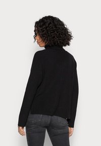Pull noir en tricot avec un col roulé, une texture côtelée et des épaules tombantes. Doté d'un dos court et d'ouvertures sur les côtés pour faciliter les mouvements.