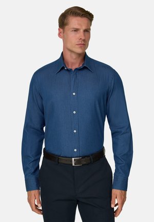 Homme portant une chemise bleue ajustée à manches longues, boutonnée, rentrée dans un pantalon foncé avec une ceinture noire sur fond blanc.
