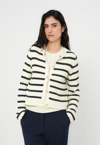 Πλεκτή ζακέτα σε κρεμ χρώμα με μαύρες και ναυτικές ρίγες, με κουμπιά μπροστά, rib κρίκο και υφή υφάσματος. Φορεμένη πάνω από κρεμ τοπ.