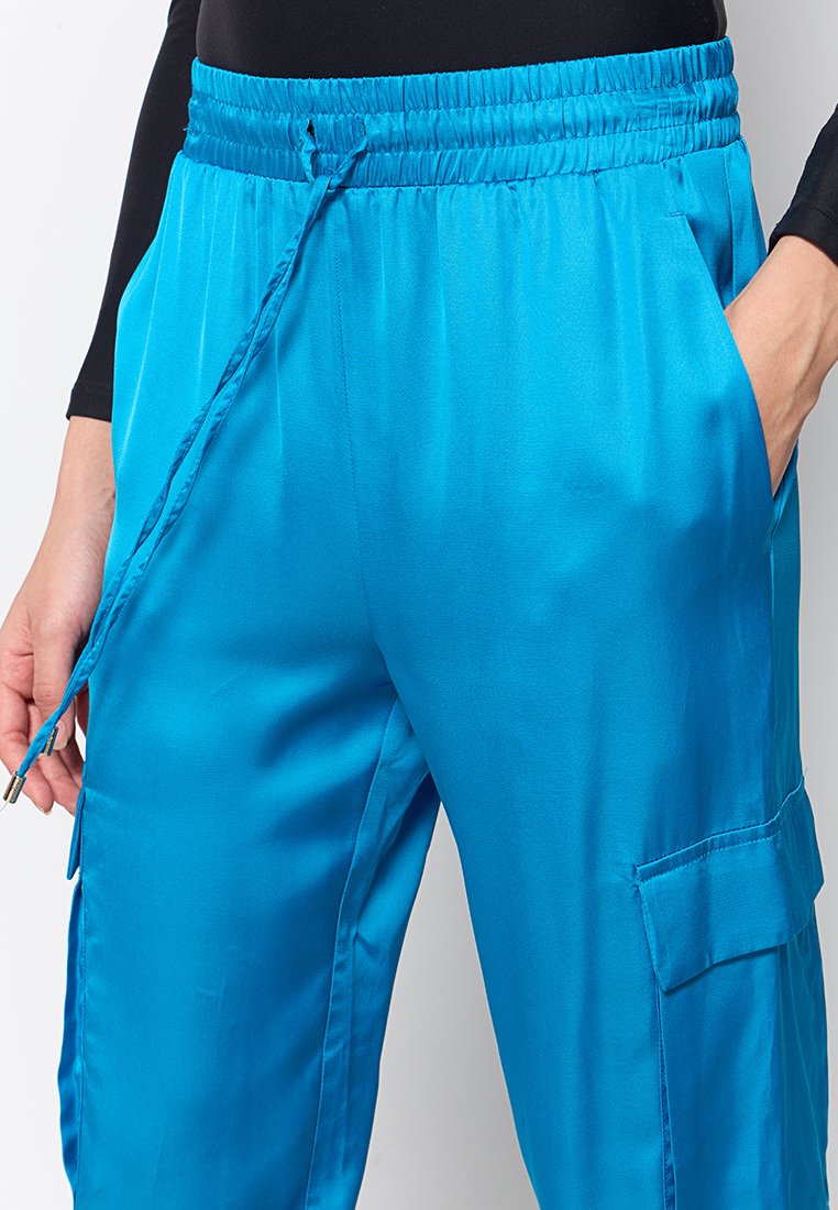 Pantalons cargo satin bleu avec une taille élastique, cordon de serrage, poches latérales et une poche cargo sur la cuisse. Texture lisse, coupe décontractée.