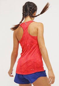 Débardeur rouge Dri-FIT avec un motif ondulé, un dos nageur et une texture lisse. Porté avec un short de sport bleu, vu de dos.