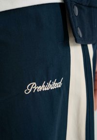 Pantaloni bleu marin cu dungi verticale crem. Prezintă cuvântul "Interzis" brodat cu un font cursiv alb. Fabricat dintr-un material neted și ușor.