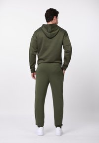 Hanorac verde închis cu fermoar, cu suprafață texturată, asortat cu pantaloni de trening similari. Pantofi sport albi completează ținuta. Vedere din spate.