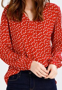 Blouse rouge à manches longues, avec un col en V, des motifs de cœurs blancs et des poignets froncés. Associée à un jean en denim foncé.