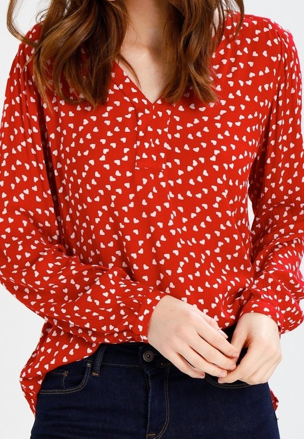Blouse rouge à manches longues, avec un col en V, des motifs de cœurs blancs et des poignets froncés. Associée à un jean en denim foncé.